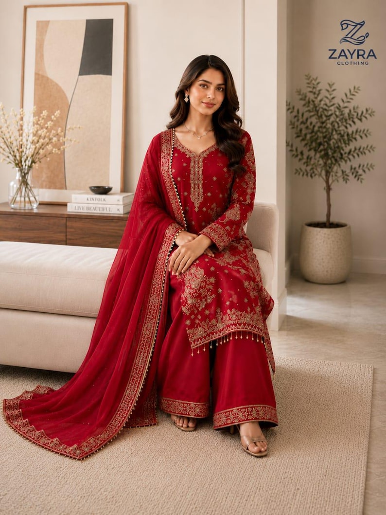 May include: A deep red long-sleeved tunic and wide-leg trousers set featuring intricate gold embroidery along the neckline, cuffs, and hem. The outfit includes a matching sheer red dupatta with a decorative gold border and small hanging beads.