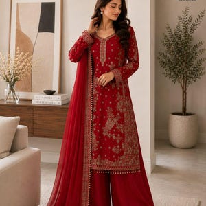May include: A deep red long-sleeved tunic and wide-leg trousers set featuring intricate gold embroidery throughout the fabric. The ensemble includes a matching sheer red dupatta with a decorative gold border and small hanging beads along the hem.