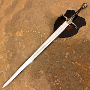 May include: A long, straight steel sword with a pointed tip and a central fuller. The hilt features a gold-toned crossguard with leaf-like details, a black grip with gold wire wrapping, and a decorative pommel, mounted on a black shield plaque.