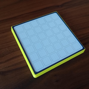 May include: A square electronic controller featuring a bright neon yellow outer frame and a dark blue border surrounding a white grid surface. The grid consists of thirty-six individual square buttons arranged in a six-by-six layout pattern.