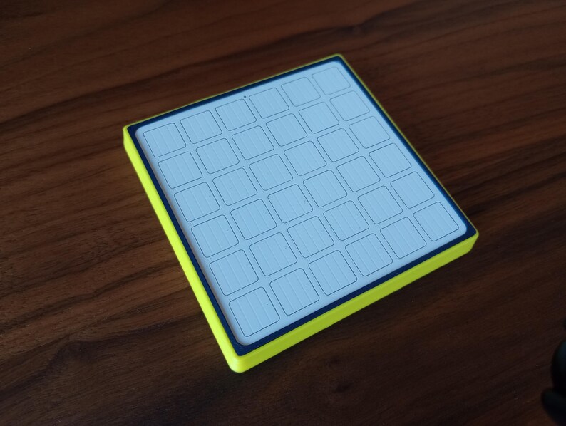 May include: A square electronic controller featuring a bright neon yellow outer frame and a dark blue border surrounding a white grid surface. The grid consists of thirty-six individual square buttons arranged in a six-by-six layout pattern.
