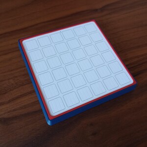 May include: A square, flat object featuring a white top surface divided into a six-by-six grid of small squares. The object has a thin red border surrounding the grid and a thick blue side edge, creating a distinct layered color profile.