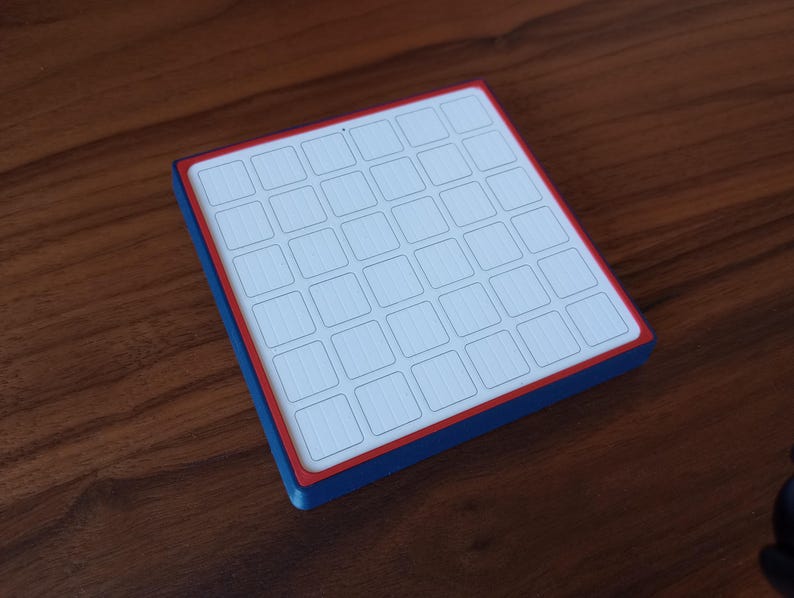 May include: A square, flat object featuring a white top surface divided into a six-by-six grid of small squares. The object has a thin red border surrounding the grid and a thick blue side edge, creating a distinct layered color profile.