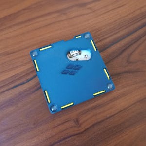 May include: A square blue electronic device housing with a matte finish, featuring yellow accent bars along the edges, four circular corner fasteners, and a central cutout revealing an internal circuit board with the text FC CE printed on it.