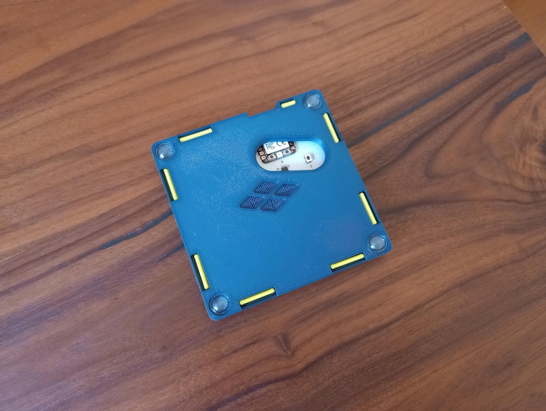 May include: A square blue electronic device housing with a matte finish, featuring yellow accent bars along the edges, four circular corner fasteners, and a central cutout revealing an internal circuit board with the text FC CE printed on it.