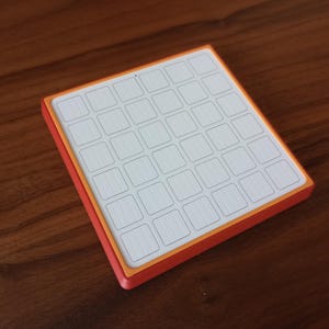 May include: A square, flat object featuring a white grid of thirty-six small, rectangular sections arranged in a six-by-six pattern. The grid is framed by a thin orange border and a thicker red outer edge, creating a distinct layered appearance.