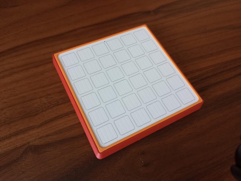 May include: A square, flat object featuring a white grid of thirty-six small, rectangular sections arranged in a six-by-six pattern. The grid is framed by a thin orange border and a thicker red outer edge, creating a distinct layered appearance.