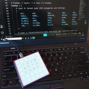 May include: A square programmable macro keypad with a white grid of keys featuring the handwritten text 'YOUR KEYS HERE' in black ink. The device is connected via a USB-C cable to a laptop displaying code with a Python logo and snake icon.