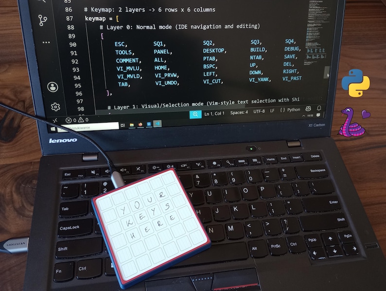 May include: A square programmable macro keypad with a white grid of keys featuring the handwritten text 'YOUR KEYS HERE' in black ink. The device is connected via a USB-C cable to a laptop displaying code with a Python logo and snake icon.