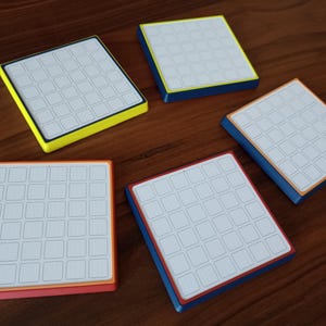 May include: Five square coasters with white surfaces featuring a grid of thirty-six small squares. Each coaster has a distinct coloured border, including yellow, blue, orange, and red, with a white top surface and a solid coloured base.