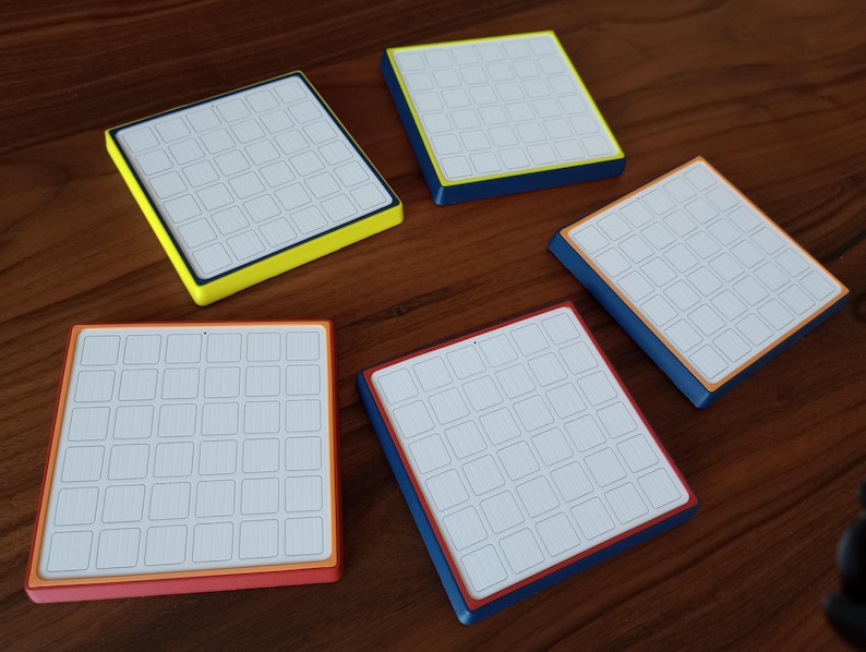 May include: Five square coasters with white surfaces featuring a grid of thirty-six small squares. Each coaster has a distinct colored border, including yellow, blue, orange, and red, with a white top surface and a solid colored base.