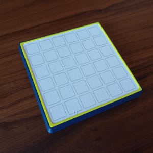 May include: A square grid controller featuring a white surface with a six-by-six array of recessed square buttons. The device is framed by a bright yellow inner border and a deep blue outer casing, creating a distinct contrast in color and form.
