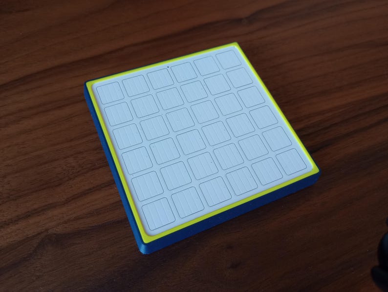 May include: A square grid controller featuring a white surface with a six-by-six array of recessed square buttons. The device is framed by a bright yellow inner border and a deep blue outer casing, creating a distinct contrast in color and form.