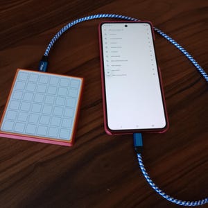 May include: A square, orange-bordered macro pad with a white grid of buttons connected by a blue and white braided cable to a red smartphone displaying a list of search results on its screen. The device is linked via a blue USB-C connector.