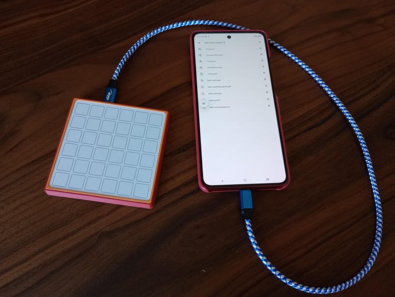 May include: A square, orange-bordered macro pad with a white grid of buttons connected by a blue and white braided cable to a red smartphone displaying a list of search results on its screen. The device is linked via a blue USB-C connector.