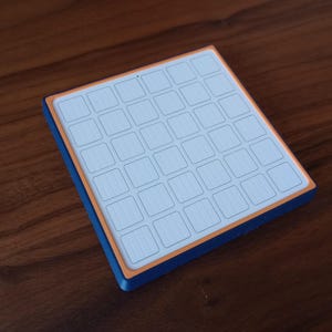 May include: A square grid-based input device featuring a white top surface with a 6x6 array of square buttons, framed by a thin orange border and a deep blue outer casing, designed for tactile interaction and precise digital control.