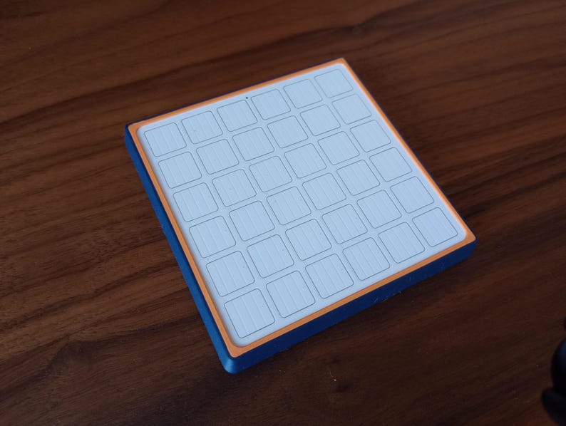 May include: A square grid-based input device featuring a white top surface with a 6x6 array of square buttons, framed by a thin orange border and a deep blue outer casing, designed for tactile interaction and precise digital control.