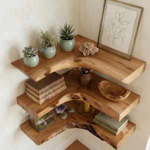 May include: Three tiered corner shelves made of natural light brown wood with live edges. The shelves hold small potted succulents, a framed line drawing, a stack of three books, a wooden bowl, a glass terrarium, and a second stack of two books.