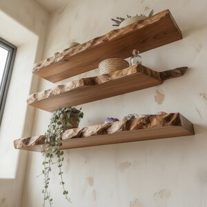 May include: Three horizontal wall-mounted shelves made of natural light brown wood with irregular, live-edge front profiles. The shelves hold various small decorative items, including a woven basket, a glass bottle, and a trailing green plant.