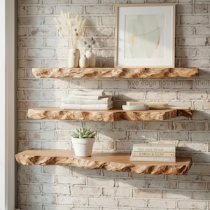 May include: Three light brown wooden floating shelves with natural live edges. The top shelf holds two ceramic vases with dried plants and a framed print. The middle shelf holds folded white towels and a soap dish. The bottom shelf holds a potted succulent and three stacked books.