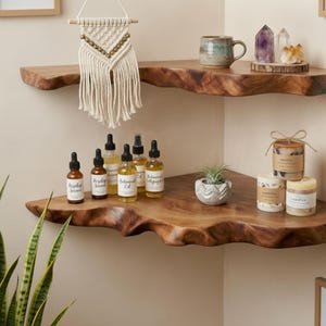 Könnte beinhalten: Zwei Eckregale aus Holz mit Naturkante an einer Wand montiert. Das obere Regal enthält einen Makramee-Wandbehang, einen Keramikbecher und zwei Kristalle. Das untere Regal zeigt fünf braune Glastropfflaschen, eine kleine Sukkulente und drei Kerzen.