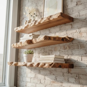 May include: Three horizontal wall-mounted shelves made of natural light brown wood with uneven, live-edge front profiles. The shelves are arranged vertically, holding small white vases with dried flowers, folded white towels, a succulent, and books.