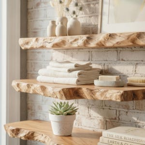 May include: Three natural live-edge wooden shelves mounted on a white brick wall. The top shelf holds three cream-colored ceramic vases. The middle shelf features a stack of folded white towels, a bar of soap in a dish, and a wooden brush. A succulent in a white pot sits on the bottom shelf.