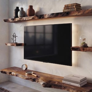 Op de afbeelding: Een aan de muur gemonteerde televisie omringd door planken van natuurlijk hout met een boomkant. De bovenste plank bevat drie vazen en boeken. De zijplanken tonen een metalen sculptuur en een glazen stolp. Een klok en loep liggen onderaan.