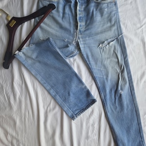 Op de afbeelding: Lichtblauwe denim jeans met distressed details, waaronder scheuren en vervaging. De jeans liggen plat op een witte ondergrond, met een houten kleerhanger aan de linkerkant. De jeans hebben een knoopsluiting.