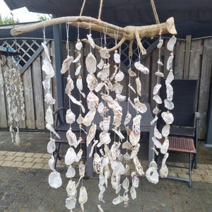 Op de afbeelding: Een handgemaakte windgong gemaakt van natuurlijke schelpen, opgehangen aan een verweerde houten tak. De schelpen zijn in verschillende vormen en maten, met tinten wit, crème en hints van paars. De gong hangt aan een natuurlijk vezeltouw, wat een kust- en bohemien esthetiek creëert.