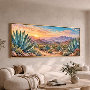 May include: A panoramic landscape wall art print in a thin gold frame depicting a desert scene at sunrise. Large green agave plants occupy the foreground, while distant mountains and a soft orange and blue sky fill the background horizon.