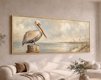 May include: A rectangular landscape painting in a thin gold frame depicting a brown and white pelican perched on a wooden piling. The scene features a calm ocean, a sandy shoreline with tall grasses, and a soft, cloudy sky in muted tones.