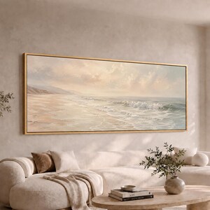 May include: A wide rectangular landscape painting in a thin gold frame depicting a serene beach scene with soft, muted tones of beige, pale blue, and white. The artwork shows gentle ocean waves rolling onto a sandy shore under a cloudy sky.