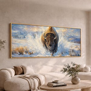 May include: A rectangular wall art print in a thin gold frame featuring a painting of a large brown bison charging forward through deep white snow. The background depicts a vast winter landscape with distant blue mountains and a cloudy sky.