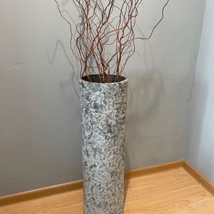 Puede incluir: Un jarrón de suelo alto y cilíndrico con un acabado texturizado y moteado en gris y blanco que recuerda a la piedra. El jarrón contiene un grupo de ramas de sauce secas, finas y sinuosas de color marrón rojizo que se extienden hacia arriba.