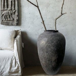 Op de afbeelding: Een grote, donker antracietkleurige vloervaas met een getextureerde, verweerde afwerking. De vaas heeft een afgerond lichaam en een smalle hals, met twee kale, lichtgekleurde houten takken die vanuit de opening omhoog steken.