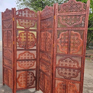 May include: A four-panel folding room divider made of reddish-brown wood. Each panel features intricate lattice work and carved floral motifs within scalloped frames. The structure is connected by metal hinges, allowing the panels to stand upright.