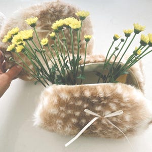 May include: Two tan and white faux fur pouches with a soft, fuzzy texture and a spotted pattern. Each pouch features a zipper closure and a small, thin white ribbon tied in a bow on the front. Bright yellow daisy-like flowers are placed inside.