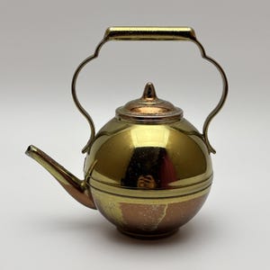 May include: A spherical metal teapot featuring a polished gold-tone upper section and a contrasting copper-toned lower base. It includes a curved handle, a pointed lid finial, and a slender spout, with visible metallic reflections on the surface.