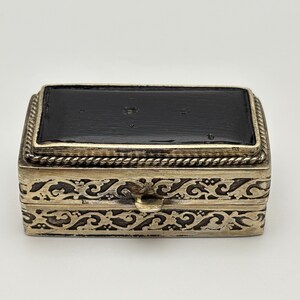 May include: A rectangular metal box featuring a smooth black lid bordered by a twisted rope-like trim. The sides of the box are decorated with an intricate, raised scrollwork pattern in a metallic finish, and a small tab is centered on the front.