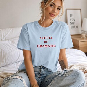 Könnte beinhalten: Hellblaues T-Shirt mit dem roten Text "A LITTLE BIT DRAMATIC". Das Model trägt eine hellblaue, zerrissene Jeans. Im Hintergrund ist ein gerahmter Druck mit dem Text "BE YOU NOT THEM" zu sehen.