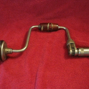 May include: A vintage metal hand brace drill featuring a U-shaped steel frame, a dark brown wooden rotating handle in the center, a round black knob at the end, and a silver metal chuck mechanism for holding drill bits securely in place.