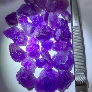 May include: A collection of raw, uncut amethyst crystals in varying sizes and shapes, displaying a deep purple hue. A pair of silver tweezers with the text "XLR1" is positioned to the right of the crystals. The crystals are on a white surface.