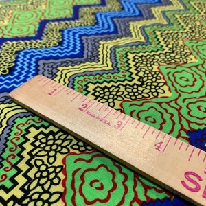 May include: A close-up of a colorful fabric with a geometric pattern. The fabric is yellow, green, blue, and black. A wooden ruler is placed on top of the fabric. The ruler is marked with inches and has the text "Made in USA" printed on it.