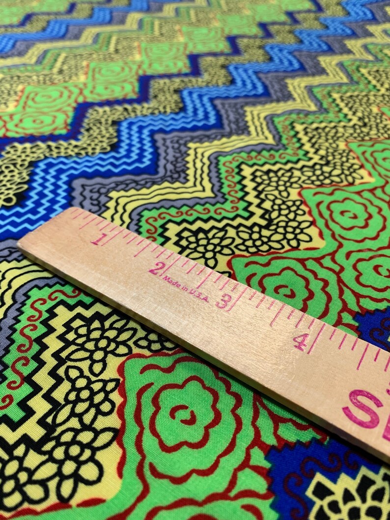 May include: A close-up of a colorful fabric with a geometric pattern. The fabric is yellow, green, blue, and black. A wooden ruler is placed on top of the fabric. The ruler is marked with inches and has the text "Made in USA" printed on it.