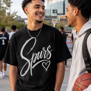 May include: Black t-shirt with the word "Spurs" in a sparkly, cursive font and a sparkly heart. Two people are smiling and talking. One person is wearing the t-shirt and a silver necklace. The other person is wearing a gray hoodie and holding a basketball.
