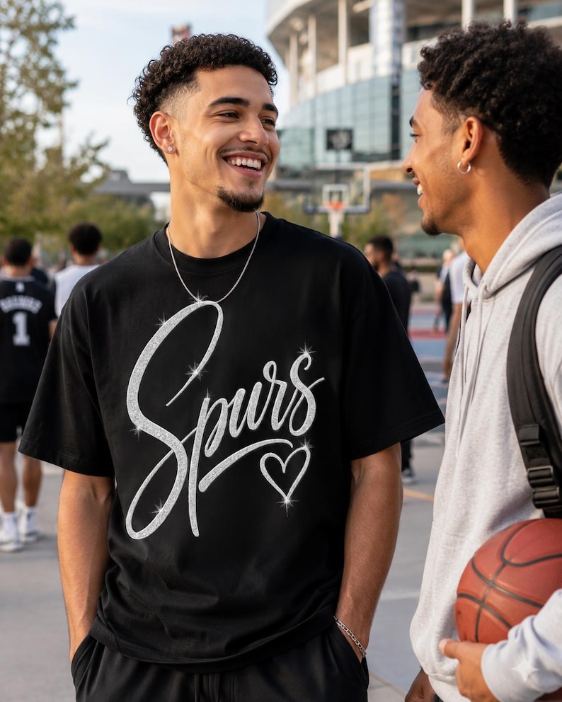 May include: Black t-shirt with the word "Spurs" in a sparkly, cursive font and a sparkly heart. Two people are smiling and talking. One person is wearing the t-shirt and a silver necklace. The other person is wearing a gray hoodie and holding a basketball.