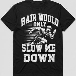 May include: A black short-sleeved t-shirt featuring a white graphic of a bald runner in motion. The text "HAIR WOULD ONLY SLOW ME DOWN" is printed in a distressed white font above and below the illustration, emphasizing a fast-paced athletic theme.