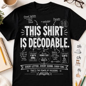 May include: A black short-sleeve t-shirt featuring white educational text and diagrams about phonics. The graphic includes the phrase THIS SHIRT IS DECODABLE along with linguistic notes on closed syllables, digraphs, and r-controlled vowels.