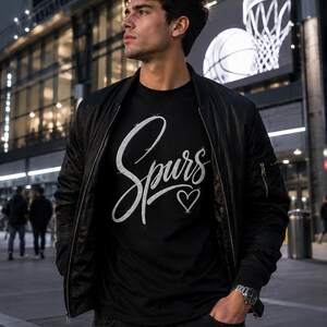 May include: A black t-shirt with the word "Spurs" in a cursive font and a heart symbol. The person is wearing a black bomber jacket and dark jeans. A basketball hoop and ball are visible in the background.